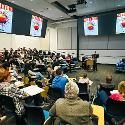 A large classroom of people sitting in desks surrounding a speaker in the middle