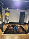 Art installation of four double sided, free hanging canvas paintings around a sectioned off square full of mulch. The artist is lying face down in the center of the mulch pit, surrounded by the paintings on each corner of the square.