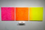 Two pieces are displayed on a gallery wall. One is a fluorescent gradient on 3 canvases from pink to orange to yellow, while the other is a pair of small folded puzzle purses sitting on a shelf below.