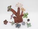 A tree-like structure made out of brown clay that has colorful ceramic flowers inserted in the stems. The piece is modular, and excess flowers lie in the surrounding space.